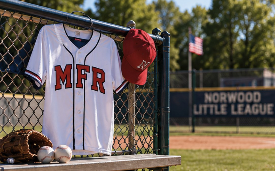 A white baseball jersey with red MFR lettering and a red cap hang on a dugout chain-link fence, with a baseball glove and two baseballs on the bench below and a Norwood Little League banner in the background.